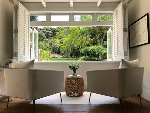 A bright, refined sitting area with two upholstered lounge chairs positioned in front of a large open window. White plantation shutters and overhead beams frame views of a lush garden, while a small woven side table with greenery sits between the chairs. Timber flooring and neutral furnishings create a calm, airy atmosphere.