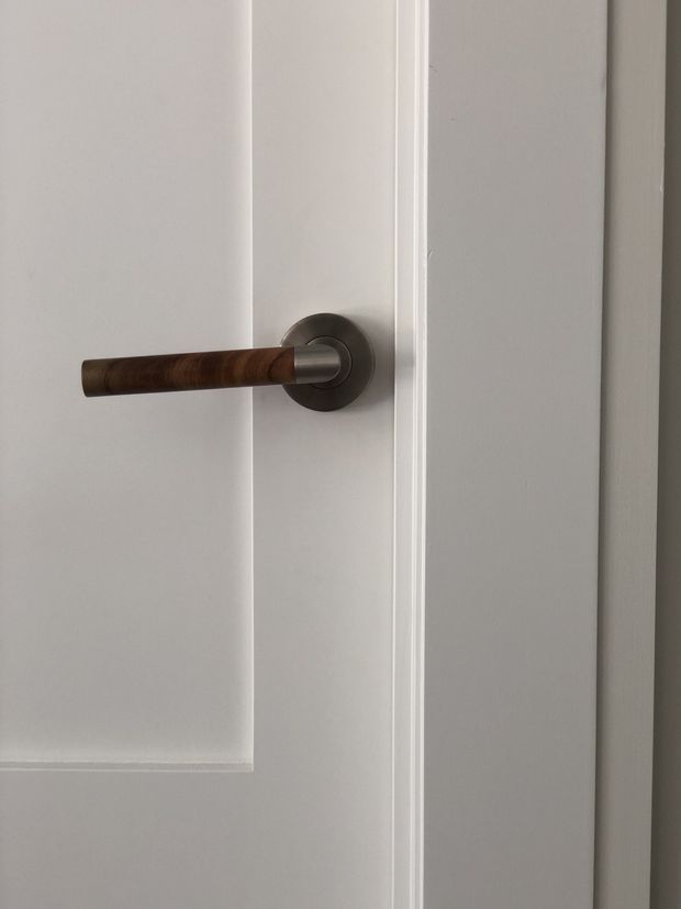 Close-up architectural detail of a white painted interior door with raised panel moulding, featuring a cylindrical wooden lever handle mounted on a dark brushed metal circular rose. The image focuses on the door hardware and trim, with no wider room context visible.