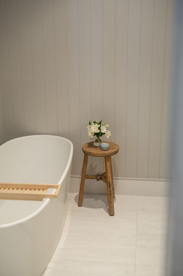 A serene, minimalist bathroom corner featuring a freestanding white bathtub with a timber bath tray, pale vertically panelled walls, light flooring, and a small rustic wooden stool holding a vase of flowers and a decorative bowl.