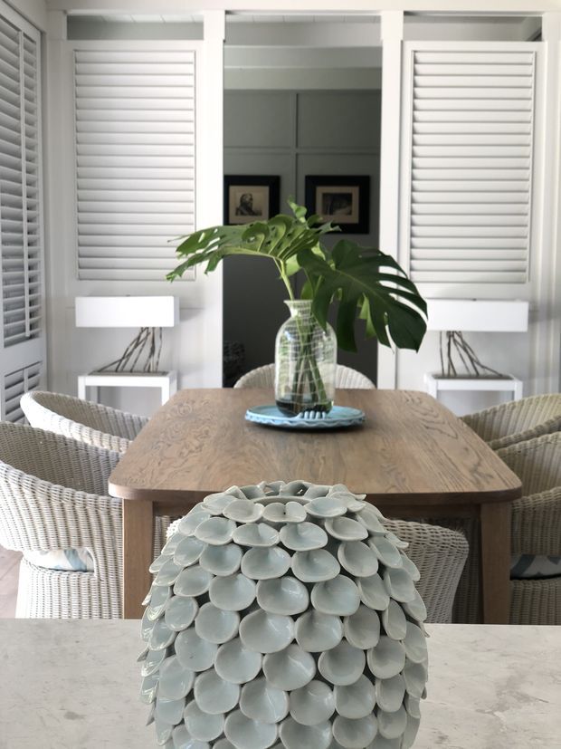 A bright dining area with a substantial natural timber dining table surrounded by woven wicker chairs. White plantation shutters cover the windows, while paired white table lamps and framed artwork sit against a dark central wall. A leafy plant in a clear glass vase forms the table centrepiece, and a pale sculptural shell-textured vase occupies the foreground on a light stone benchtop. The room has a relaxed, coastal-inspired feel with layered natural textures and neutral tones.