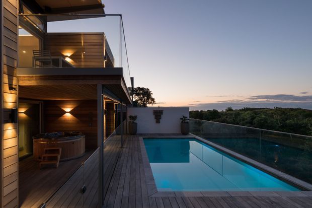 Contemporary multi-level residential exterior at dusk, featuring warm timber cladding and soffits, expansive glazing, a timber deck, and a rectangular swimming pool enclosed by frameless glass balustrades. The pool terrace overlooks a wooded landscape, with integrated wall and soffit lighting creating a warm, refined atmosphere.