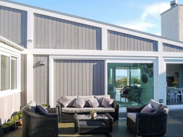 A bright contemporary courtyard-style exterior with a covered outdoor seating area in front of a single-storey building. The facade combines pale vertical weatherboard cladding, white trim, large glazed sliding doors, and a simple sloping roofline. Wicker outdoor sofas and armchairs surround a low table on a dark deck or paved surface, with a dining area visible through the glazing.