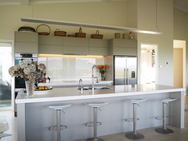 Bright, spacious contemporary kitchen with a large white island and integrated breakfast bar seating for four. The kitchen features pale grey cabinetry, a glossy light splashback, stainless-steel appliances including a large refrigerator, twin sinks with modern mixer taps, and open shelving displaying woven baskets. A long linear pendant light hangs above the island, while neutral finishes and fresh flowers create a clean, airy atmosphere.