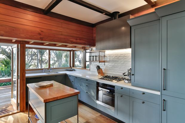 A warm, characterful kitchen with blue-grey shaker cabinetry, a timber island and timber flooring. The room features extensive glazing overlooking greenery, white textured tile splashback, stainless steel appliances, dark exposed ceiling beams, and a richly stained timber wall and window framing.