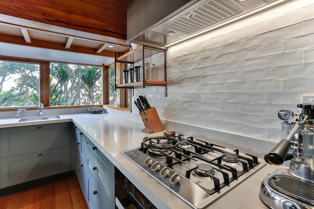 A bright, compact kitchen viewed from the work area, featuring a long light benchtop, grey-blue shaker-style cabinetry, a large gas cooktop, stainless-steel rangehood, tiled backsplash, timber flooring and a timber-lined ceiling. A window overlooks leafy vegetation, while open shelving and countertop appliances add functional detail.