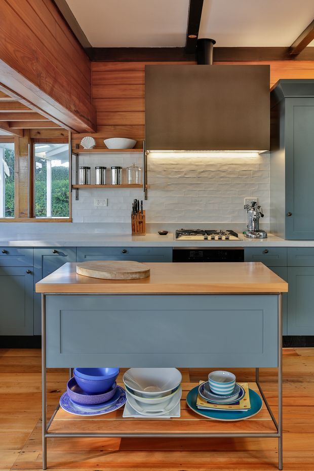 A warm, compact kitchen with blue-gray shaker cabinetry, a central island with a timber benchtop and open lower shelving, white textured tile backsplash, timber wall panelling and flooring, and dark exposed ceiling beams. A gas cooktop, stainless espresso machine, open shelf storage, and colourful crockery are visible. Large windows bring in natural light and overlook greenery.