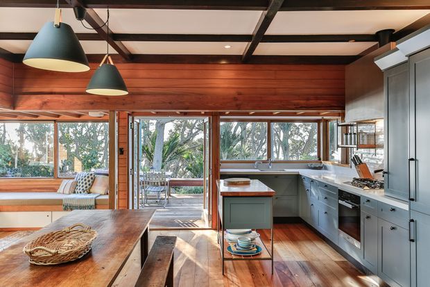 Warm, timber-lined kitchen and dining area with extensive glazing overlooking trees. The room features dark blue-grey cabinetry, timber benchtops and flooring, exposed dark ceiling beams, pendant lights, and a built-in window seat adjoining an outdoor deck.