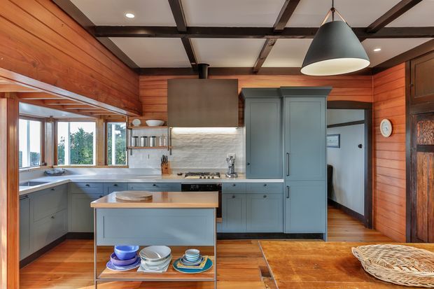 A warm, compact kitchen with blue-grey cabinetry, a central island, timber flooring, and extensive horizontal timber wall panelling. The room features a white textured tile splashback, timber-framed windows, exposed dark ceiling beams, and a large black pendant light. Open shelving and a freestanding display trolley add practical storage, while a doorway leads to a hallway.