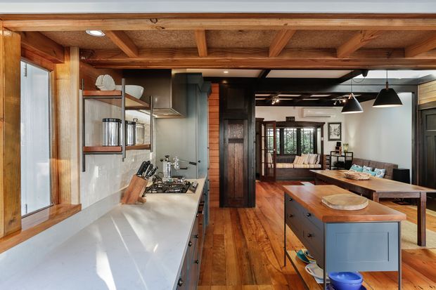 A warm, open-plan kitchen and living area with extensive timber framing and exposed ceiling beams. The kitchen features a long pale benchtop, dark cabinetry, stainless steel appliances, open shelving, and a central island with a timber benchtop. Black pendant lights hang over the island, while the adjoining living and dining area is visible beyond.