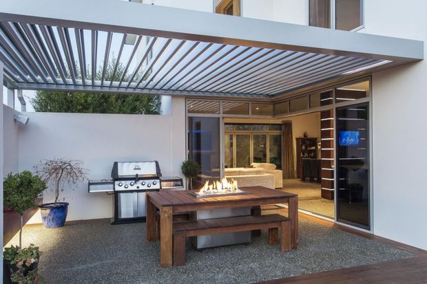 Contemporary covered courtyard entertaining area with a timber dining table, built-in linear fire feature, barbecue grill, and large glazed doors opening to an interior living space. The space is enclosed by white rendered walls and shaded by a modern timber-slatted pergola.