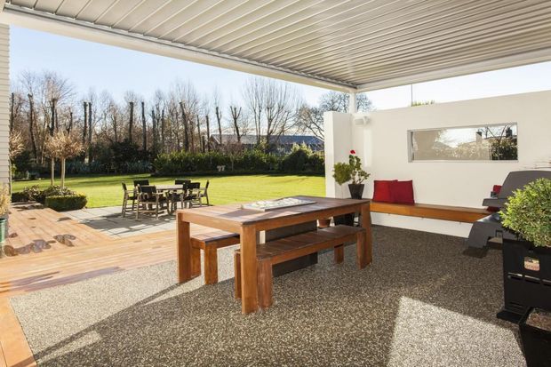 Covered outdoor entertaining area opening onto a timber deck and lawn. The space features a long solid timber dining table with benches, a built-in white rendered bench seat with red cushions, a textured paved floor, a horizontally slatted ceiling, and a large rectangular opening framing the garden.