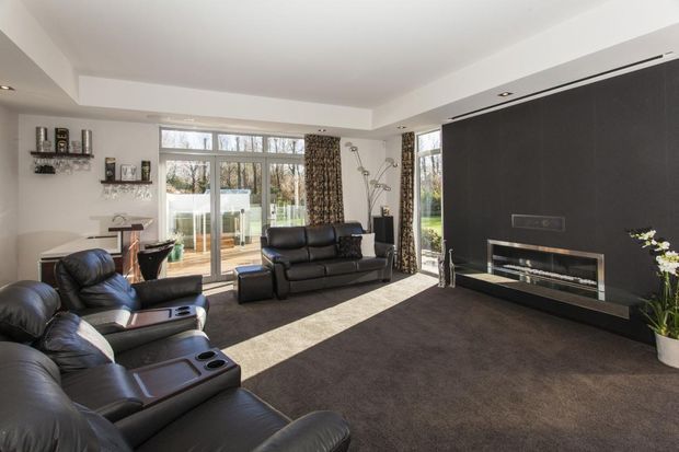 Contemporary living room with black leather reclining sofas, a large dark feature wall incorporating a sleek linear fireplace and television, floor-to-ceiling glazing, and neutral carpet. Natural light creates a bright, comfortable media-focused space.