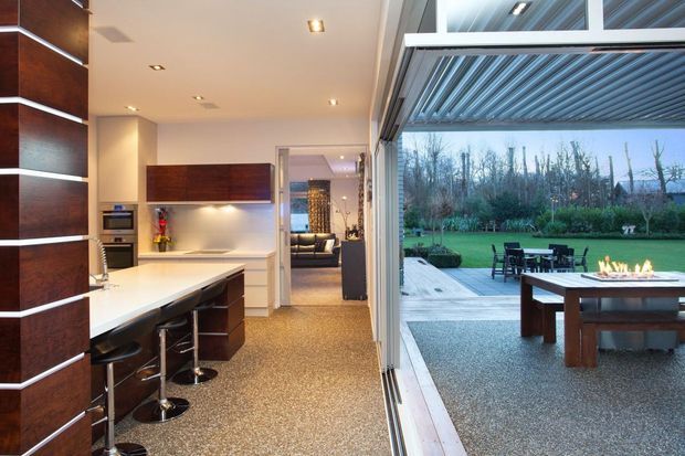 Contemporary open-plan kitchen with dark timber cabinetry, a pale stone island, integrated appliances and bar seating. The kitchen opens through fully glazed, sliding doors to a paved outdoor entertaining area with a dining table and linear fire feature, overlooking landscaped grounds. Recessed ceiling lighting and a louvred outdoor roof create a refined indoor-outdoor connection.