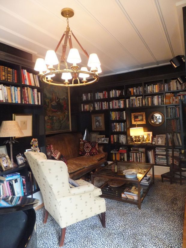 A richly layered traditional library or study with floor-to-ceiling dark built-in bookshelves, a brown leather sofa, patterned upholstered armchair, glass-topped coffee table, framed artwork, and a large brass chandelier. The room has a warm, intimate, moody atmosphere with a highly patterned rug and a white panelled ceiling.
