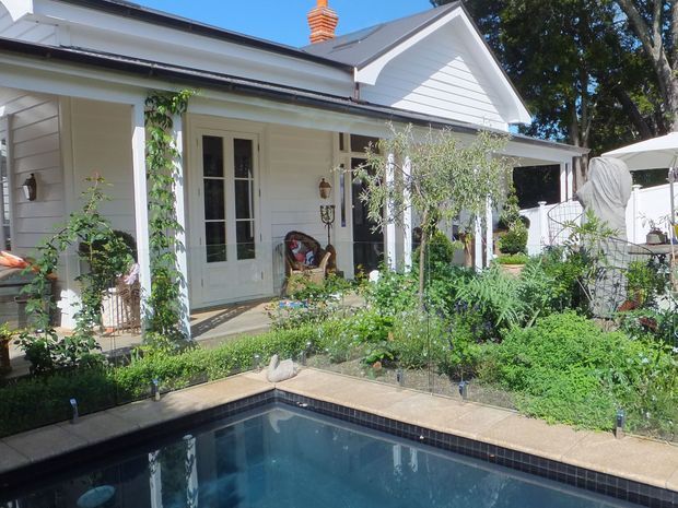 A bright exterior view of a white weatherboard house with a gabled roof, covered front veranda, French doors and landscaped garden beds. A swimming pool occupies the foreground, with outdoor seating and umbrellas visible to the right. Mature trees surround the property.