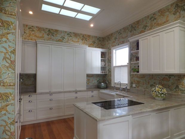 A bright, traditionally styled kitchen with extensive white paneled cabinetry, a large central island with an integrated black cooktop, pale stone benchtops, patterned botanical wallpaper, timber flooring, and a ceiling skylight. A farmhouse-style sink sits beneath a window, with open shelving displaying decorative items.