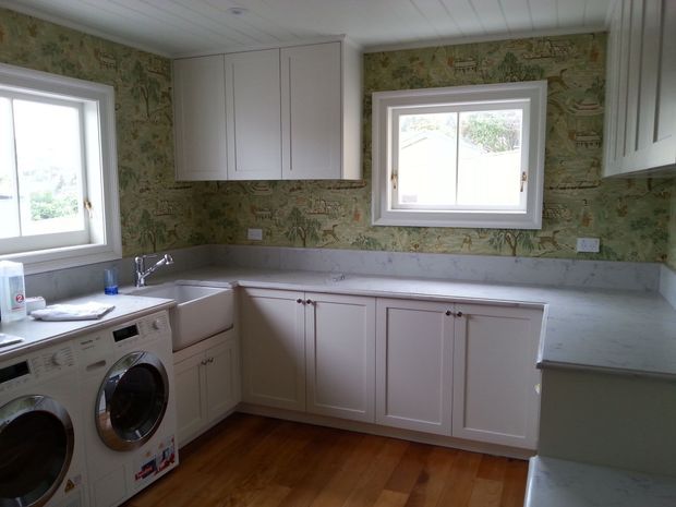 A bright, traditional-style laundry room with extensive pale cabinetry, grey benchtops, a farmhouse-style sink, front-loading washing machines, timber flooring, and a distinctive vintage map-patterned wallpaper backsplash. White-framed windows provide natural light.
