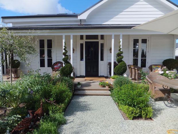 Bright front exterior of a single-storey home with a white horizontal weatherboard facade, gabled roof, covered entry porch, dark glazed front door, and symmetrical tall windows. A gravel path leads through a neatly landscaped garden with clipped shrubs, topiary, and outdoor wicker seating.