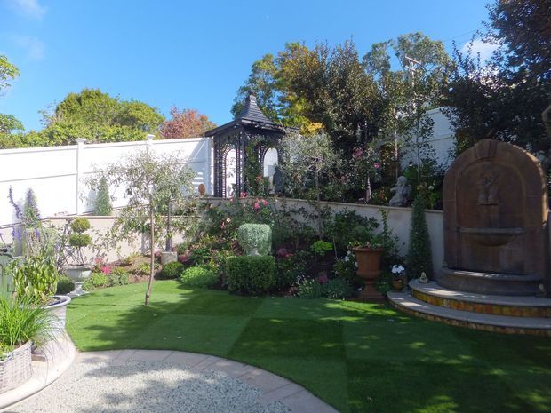 A landscaped courtyard garden with clipped hedges, flowering plants, artificial turf, curved pale gravel paving, and a decorative arched metal garden structure. A large rustic stone or timber-toned wall fountain stands on a raised tiled platform at the right, with high rendered boundary walls and mature trees creating a secluded outdoor setting.