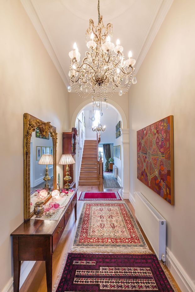 A narrow, high-ceilinged traditional entry hall with ornate white mouldings, a large crystal chandelier, an arched opening leading to a timber staircase, and patterned runners across the floor. A dark timber console with an ornate gold-framed mirror and decorative lamps lines the left wall, while colourful framed artwork hangs on the right. The space has a richly decorated, formal and eclectic character.