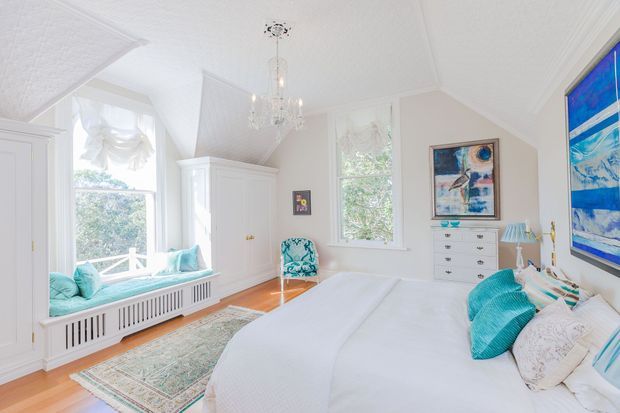 Bright, airy bedroom with white walls and ceiling, a large upholstered bed, turquoise and white bedding, built-in window seating, white cabinetry, a patterned rug, timber flooring, and a crystal pendant light. Large windows provide abundant natural light, while colourful coastal-style artwork adds visual interest.