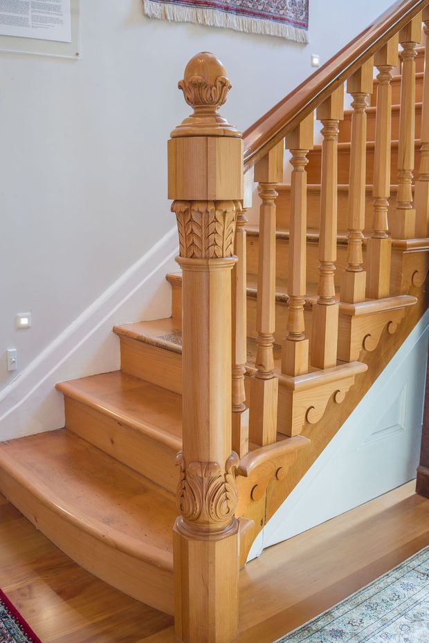 Close-up interior view of an ornate traditional timber staircase. The staircase features polished honey-toned wood treads, turned balusters, carved newel posts with acanthus-style detailing, decorative carved apron panels, and a patterned runner visible at the lower edge. Pale painted walls and white trim surround the stair, with a light blue painted area beneath the landing.