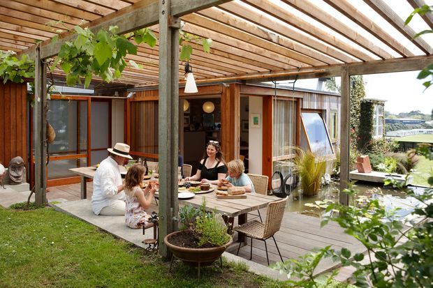 A warm outdoor dining and entertaining area beneath a timber pergola, with a timber deck, dining table, built-in garden planting, and climbing vines. The space connects directly to a timber-clad dwelling with large glazed openings and overlooks a landscaped garden and neighbouring landscape. Several people are gathered around the table, creating an inviting residential courtyard atmosphere.