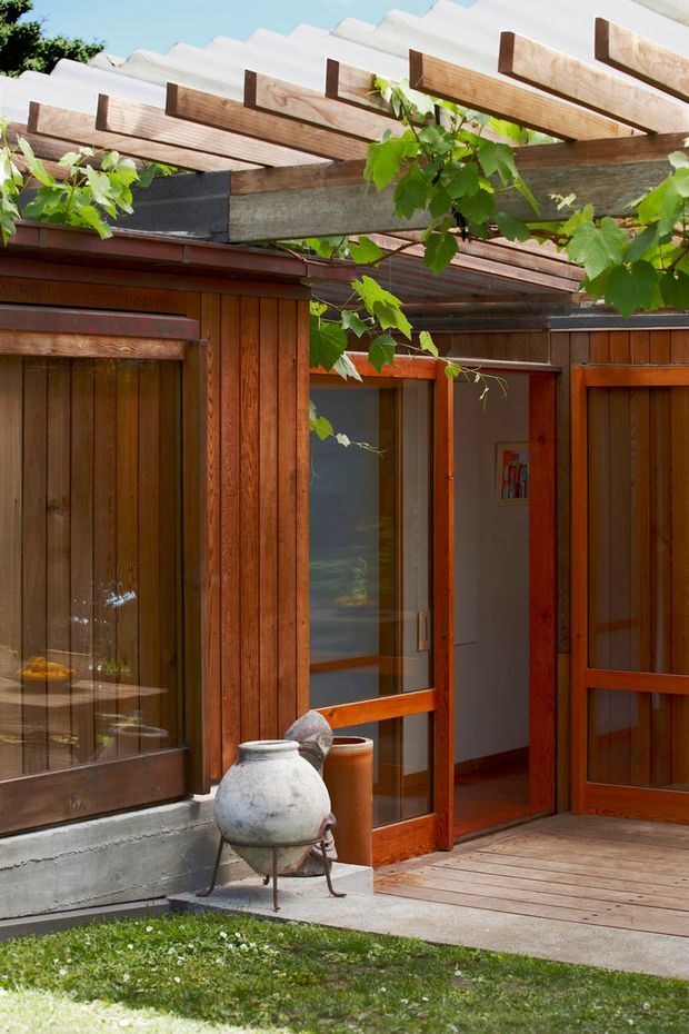 A warm timber-clad courtyard or entry exterior with glass-panelled wooden doors and windows, a timber pergola supporting leafy vines, and a small timber deck beside a planted lawn. The facade combines rich reddish-brown wood, grey structural beams, and a corrugated roof edge, creating a rustic, organic outdoor setting.