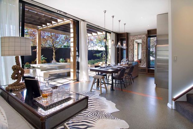 Open-plan contemporary kitchen and dining area with extensive floor-to-ceiling glazing opening onto a covered outdoor entertaining space. The room features dark timber cabinetry, a long island or dining counter with stools, a formal dining table, polished concrete-style flooring, pendant lights, and a refined sitting area with a console and zebra-pattern rug. A stairway is visible at right.