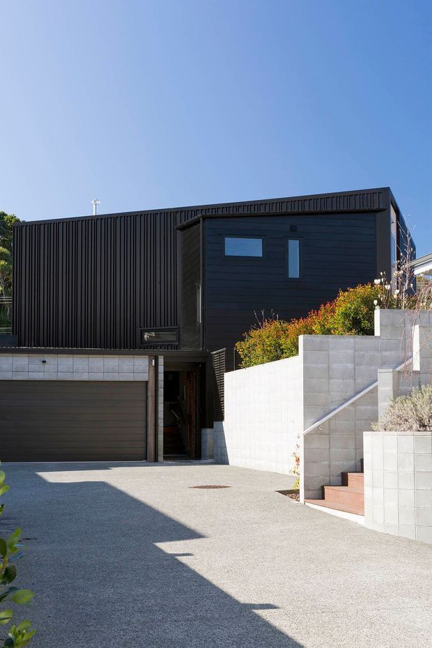 Exterior view of a contemporary multi-level residence with a dark charcoal facade, vertical slatted cladding, horizontal timber-like wall siding, a garage, and a recessed entry. Pale concrete block retaining walls and stairs step up through landscaped planting. The image is captured in bright daylight against a clear blue sky.