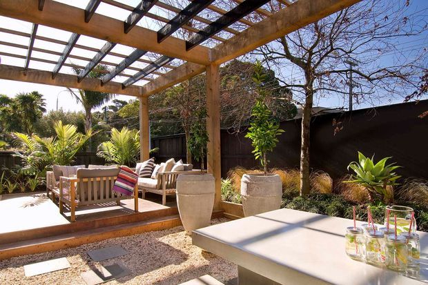 A landscaped outdoor entertaining area with a timber pergola and translucent roof over a raised deck. The deck contains timber lounge seating with striped cushions, while a pale paved and gravel courtyard surrounds a large outdoor table with drinks. Tropical planting, rendered planters, ornamental grasses, and a dark boundary fence create a warm contemporary garden setting.