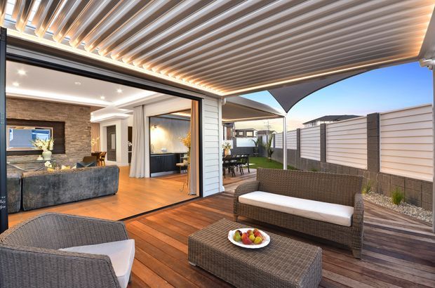 A contemporary indoor-outdoor living area opening onto a timber deck. The covered alfresco space has a slatted roof with integrated lighting, woven outdoor lounge furniture, a coffee table, and a white-cushioned bench. Through the wide glazed opening is a warm living room with a sectional sofa, timber flooring, recessed lighting, and a textured stone feature wall. A landscaped courtyard and privacy fencing are visible beyond.