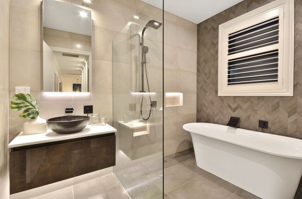 Contemporary bathroom with a freestanding white bathtub, glass-enclosed shower, floating dark timber vanity, vessel basin, large mirror, and neutral beige tiled surfaces. A textured chevron-pattern feature wall and louvred window add visual interest.