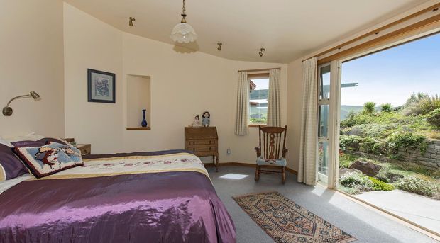 A bright bedroom with a large bed dressed in a purple satin coverlet and patterned pillows, a timber dresser, upholstered armchair, framed artwork, and a traditional pendant light. Full-height glazed doors open to a landscaped garden and bring in abundant natural light.