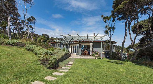 Exterior view of a low-rise contemporary coastal-style house set within a landscaped garden, with extensive glazing, a pergola-like exposed roof structure, and a stepping-stone path leading to a covered patio entrance. Wind-shaped trees and open lawn surround the dwelling.