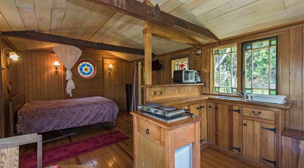 Rustic timber-lined interior combining a compact kitchen with an adjacent bedroom. The kitchen has warm wood cabinetry, a central island with cooktop, a freestanding stove, and a sink beneath leaded-style windows. The bedroom includes a timber bed, colourful patterned rug, decorative wall lighting, and a small stained-glass feature. Exposed timber beams and plank ceilings contribute to a cosy cabin-like character.