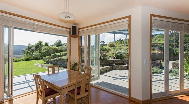 Bright dining area with a timber dining table and chairs beside extensive floor-to-ceiling glazed doors and windows. The room opens visually to a landscaped garden and paved outdoor terrace, with warm timber flooring, white walls, timber-toned window trims, and woven blinds.