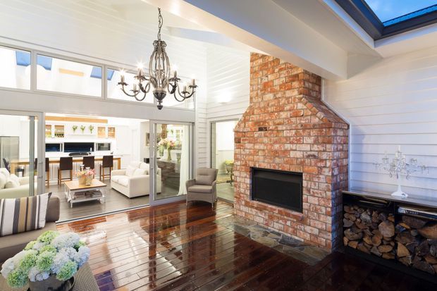 Bright open-plan living room with a large exposed red-brick fireplace, dark polished timber flooring, white horizontal wall panelling, a traditional chandelier, and clerestory glazing. Sliding glass doors open toward a dining and kitchen area, while a skylight brings in additional daylight.