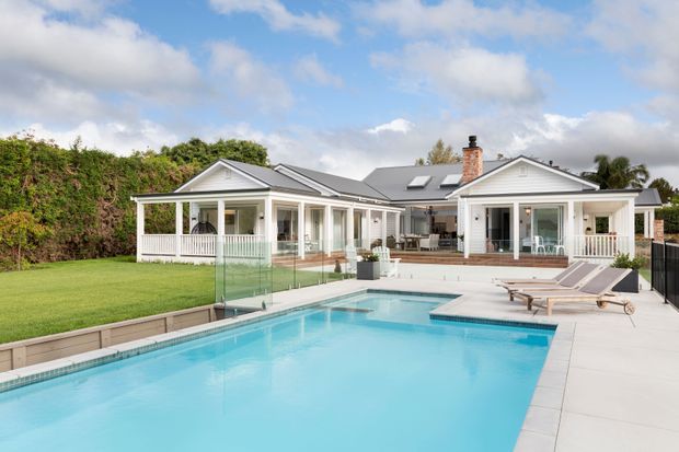 A bright rear exterior view of a large single-storey home with white weatherboard-clad gabled wings, covered verandas, extensive glazing and a central roof section. A substantial turquoise swimming pool with a glass balustrade and pale paved surround dominates the foreground, with manicured lawn and hedging behind.