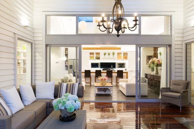 Bright, refined open-plan living area viewed from a covered or enclosed sitting space, with white horizontal wall panelling, polished dark timber flooring, large grey-framed glazed openings, and a dramatic traditional chandelier. A large sectional sofa and armchairs frame the foreground, while a dining area and kitchen are visible beyond.