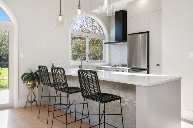 Bright contemporary kitchen with a large white island and waterfall edge, integrated seating for three, white cabinetry, stainless-steel refrigerator and cooktop, and a patterned stone-look island face. Arched windows and a glazed door bring in abundant natural light, while pendant lights and woven black bar stools add contrast.