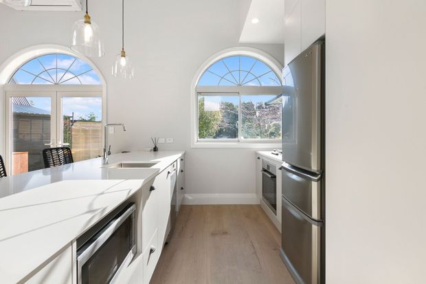Bright contemporary kitchen with white cabinetry, a large white island with integrated appliances and sink, stainless-steel refrigerator and oven, pale timber flooring, and arched windows and doors bringing in abundant natural light. Pendant lights hang above the island, and a dining area is visible near the outdoor opening.