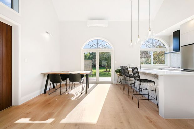 Bright open-plan kitchen and dining area with light timber flooring, white walls and cabinetry, a white island with black bar stools, and a simple timber dining table. Tall arched glazed doors and windows bring in abundant sunlight, creating a clean contemporary interior.