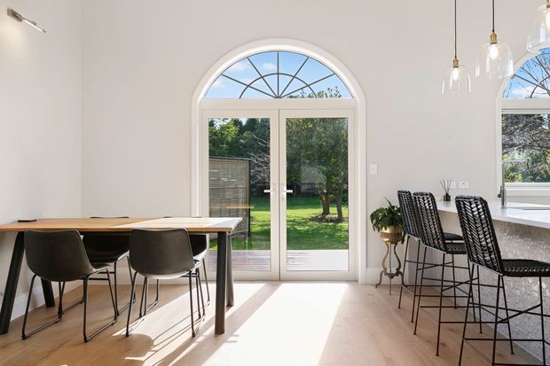 Bright open-plan dining and kitchen area with pale timber flooring, white walls, and a large arched glazed French door opening to a leafy garden. A dark timber dining table with black chairs occupies the left side, while a stone kitchen island with woven black bar stools and pendant glass lights is visible on the right. The space has abundant natural light and a clean, contemporary feel.