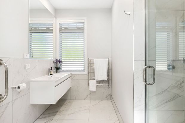 Bright contemporary bathroom with white marble-look wall and floor tiles, a floating white vanity with integrated basin, frameless glass shower enclosure, chrome fixtures, and a heated towel rail. Shuttered windows provide natural light, creating a clean, airy feel.