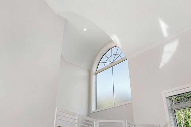 Bright white interior architectural detail featuring a tall arched window with decorative radial muntins and frosted lower glazing. The ceiling curves into a shallow barrel-vault form with white lining boards, while partial white balustrade or stair railing is visible below and a louvred window appears at right. Strong daylight creates a clean, airy atmosphere.
