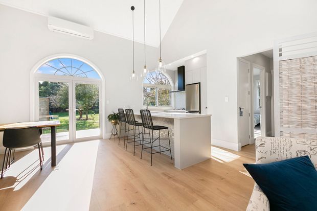 Bright open-plan kitchen and living area with white walls, light timber flooring, a white island with four dark bar stools, arched glazed doors and windows, pendant lights, and partial dining and lounge furniture. Strong natural light creates an airy contemporary interior.