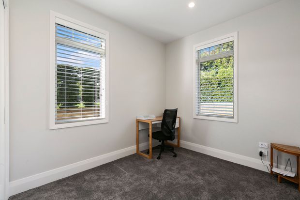 A small, bright home office with two windows fitted with white plantation-style blinds, pale neutral walls, grey carpet, and simple timber furniture including a compact desk and office chair.