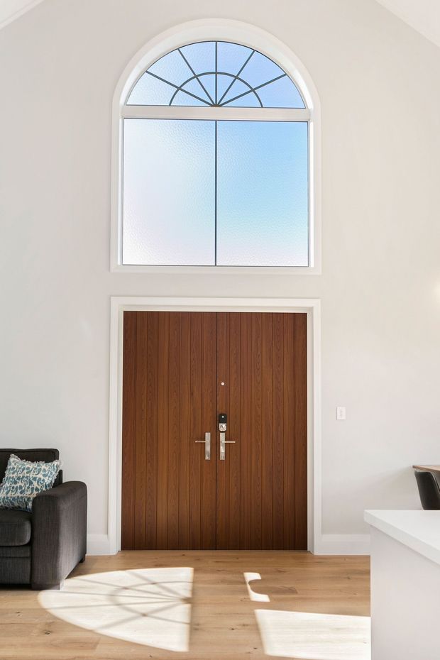 Bright residential entryway with a tall double wooden front door featuring vertical timber panelling and brushed metal hardware. Above it is a large arched frosted-glass fanlight window with decorative radial muntins. The space has white walls and trim, light timber flooring, and partial views of a dark armchair and white kitchen or console joinery.