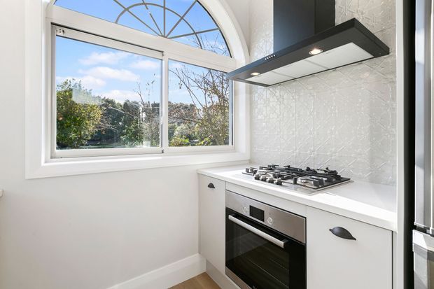 Bright compact kitchen with white cabinetry and benchtops, a stainless-steel built-in oven and gas cooktop, textured white tiled splashback, and a black integrated rangehood. A large white-framed arched window provides abundant natural light and views of greenery.